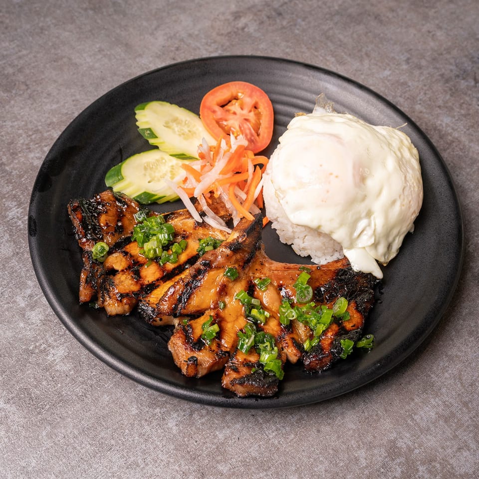 Filipino grilled pork chops with rice, fried egg, sliced cucumbers, tomatoes, and pickled vegetables.