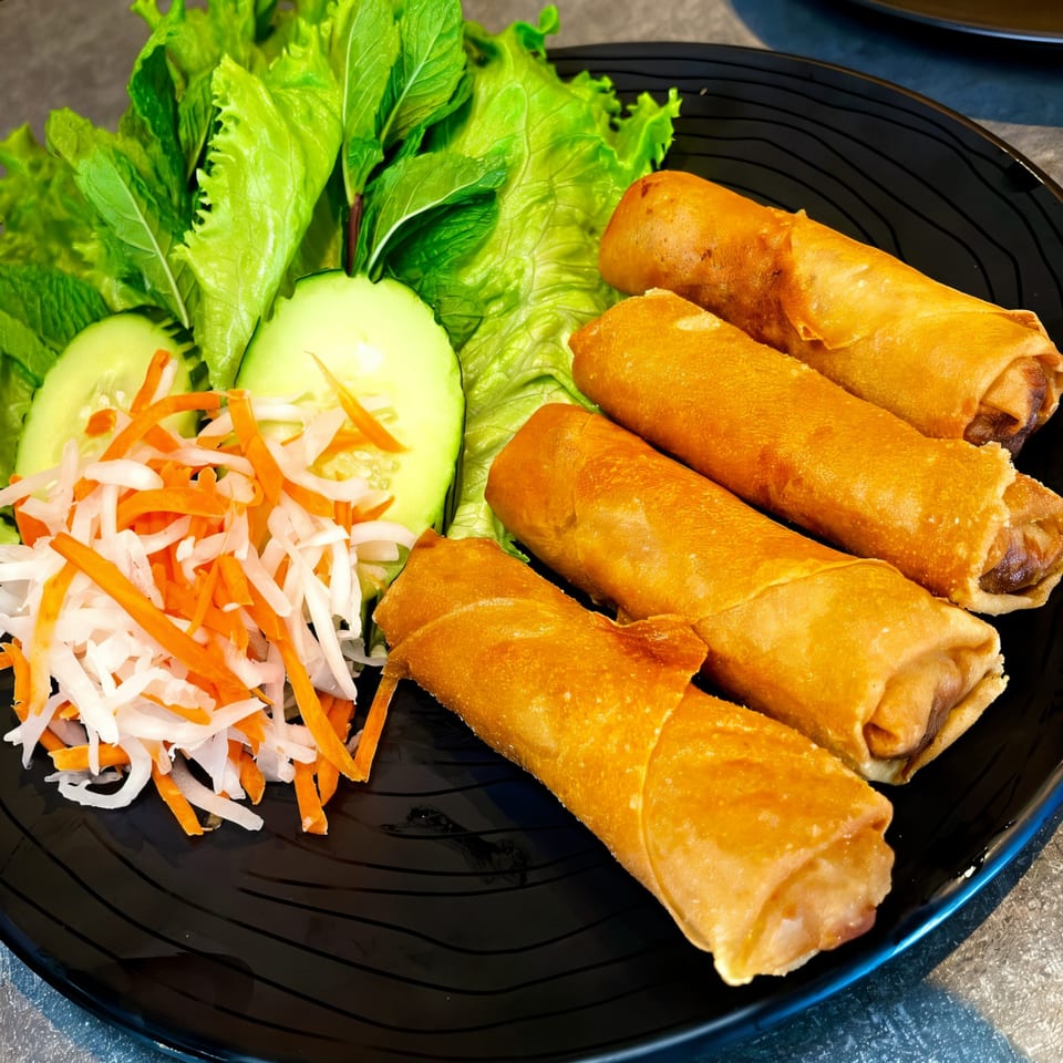 Four golden-brown Filipino lumpia spring rolls on a black plate with fresh lettuce, cucumber, and pickled vegetables.