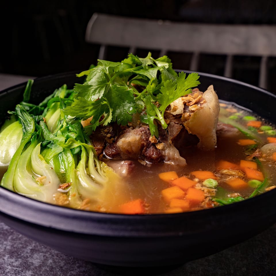 A black bowl of Mang Felix Kitchen Rainbow's Filipino beef nilaga soup with bok choy, carrots, and cilantro.